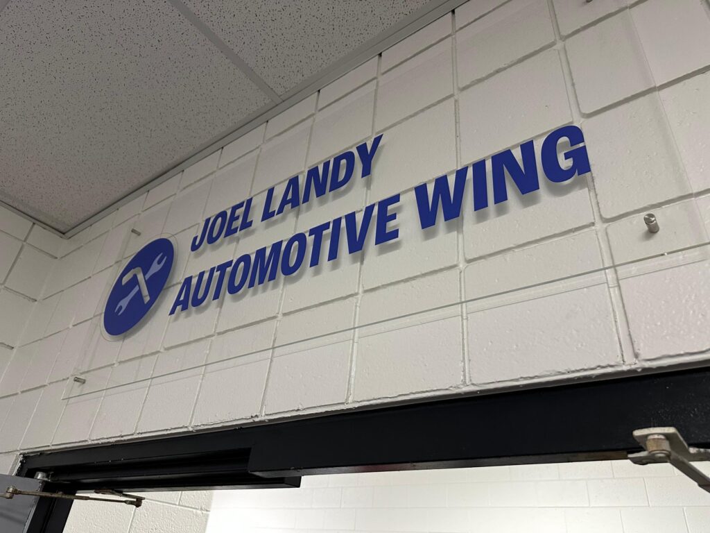A sign reading "Joel Landy Automotive Wing," featuring a hammer and wrench icon, is mounted on a white tiled wall near the ceiling. This space is part of a donation to the Detroit school's auto training center, fostering future automotive expertise.